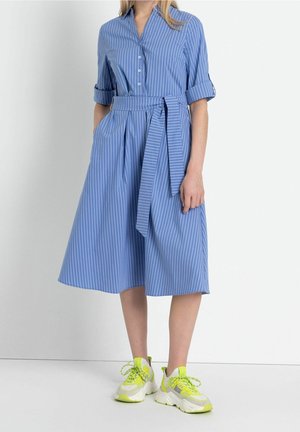 Kvinde iført en blå, hvidstribet midi kjole med knapper, opkrogede ærmer og bundet bælte i taljen, kombineret med hvide og neon gule sneakers.