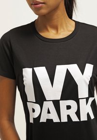 Camiseta de algodón negra con un gran gráfico blanco de "IVY PARK" en el frente. Mangas cortas, cuello redondo y un diseño ajustado.