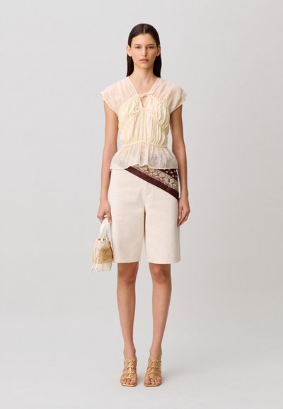 Giovane donna che indossa una camicetta in tessuto trasparente color crema, pantaloncini beige fino al ginocchio con una fascia marrone a fantasia, tacchi gialli con lacci e che tiene una borsa beige.