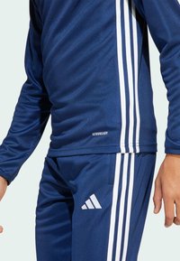 Marineblauwe sporttracksuit gemaakt van gladde stof, met witte strepen op de mouwen en broek. Inclusief "AEROREADY" branding en logo.