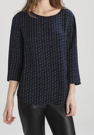 Femme portant un haut bleu marine à manches longues avec de petits motifs à pois blancs et un pantalon en cuir noir, debout sur un fond uni.