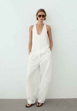 Femme portant un haut blanc sans manches à décolleté plongeant en V, un pantalon blanc à jambes larges, des lunettes de soleil marron et des tongs noires, debout devant un mur blanc uni.