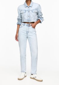 Veste courte en denim bleu clair avec deux poches avant et fermeture à boutons, associée à un jean taille haute bleu clair et des baskets blanches.
