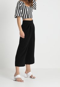 Černé kalhoty culottes s vysokým pasem v kombinaci s černobílým pruhovaným krátkým topem a bílými sandály na širokém podpatku.