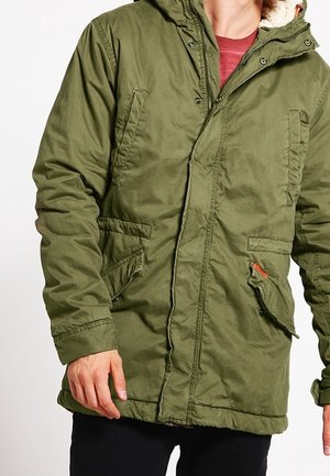 Homme portant une parka vert olive avec une capuche bordée de fourrure et plusieurs poches avant, associée à une chemise rouge et un pantalon noir.