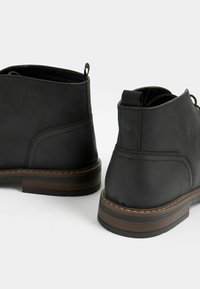 Botas negras de cuero hasta el tobillo con una textura suave, puntera redonda y detalles cosidos. El tacón presenta un acento de madera en contraste.
