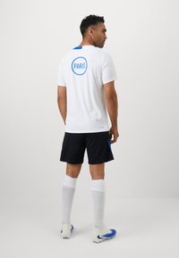 Kortærmet hvid trøje med blåt "PARIS" logo på ryggen, sorte shorts, hvide knæhøje sokker og blå fodboldstøvler, der viser atletisk design og pasform.