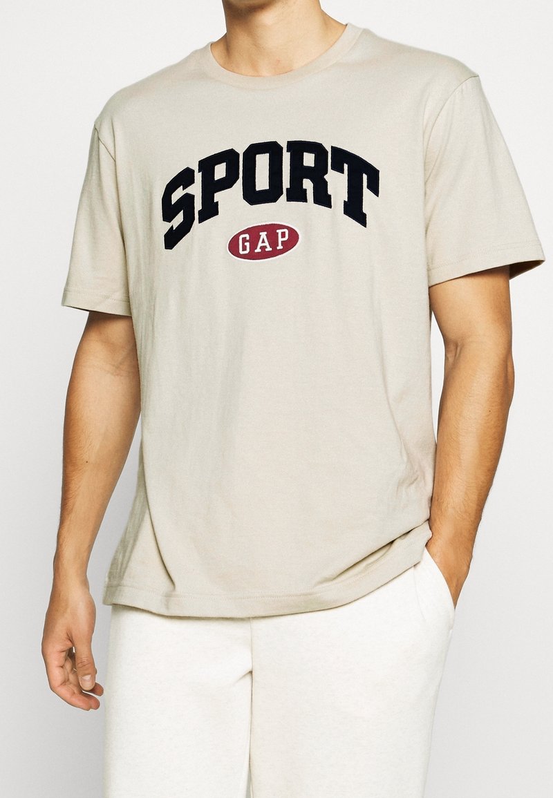 Homme portant un t-shirt beige à manches courtes avec le logo "SPORT GAP" en noir et rouge, associé à un pantalon blanc, main dans la poche.