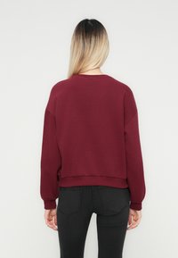 Kvinna med rakt blont hår som bär en långärmad maroon tröja och svarta jeans, stående vänd mot en enkel vit bakgrund.
