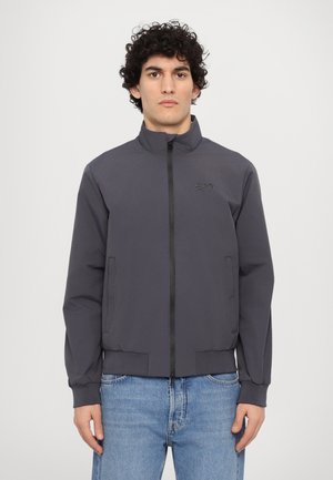 Giacca leggera grigia con collo alto e zip frontale. Presenta polsini elasticizzati e tasche laterali, abbinata a jeans blu.