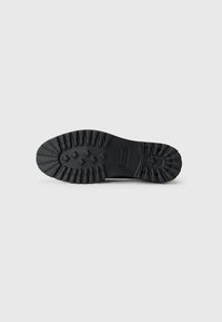 Suela de goma negra con un patrón texturizado, que presenta ranuras profundas y crestas para mayor tracción. Perfil plano y diseño de punta redondeada.