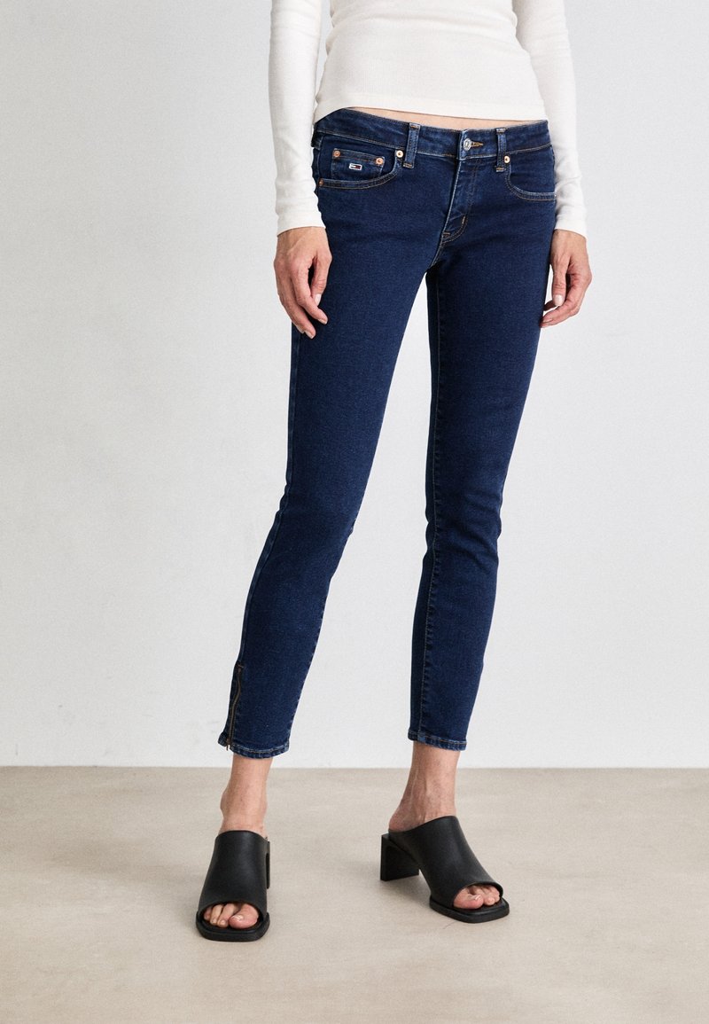 Jeans skinny de mezclilla azul oscuro con cinco bolsillos, costuras naranjas y una pequeña abertura en el tobillo. Combinados con tacones de bloque negros y abiertos por delante.