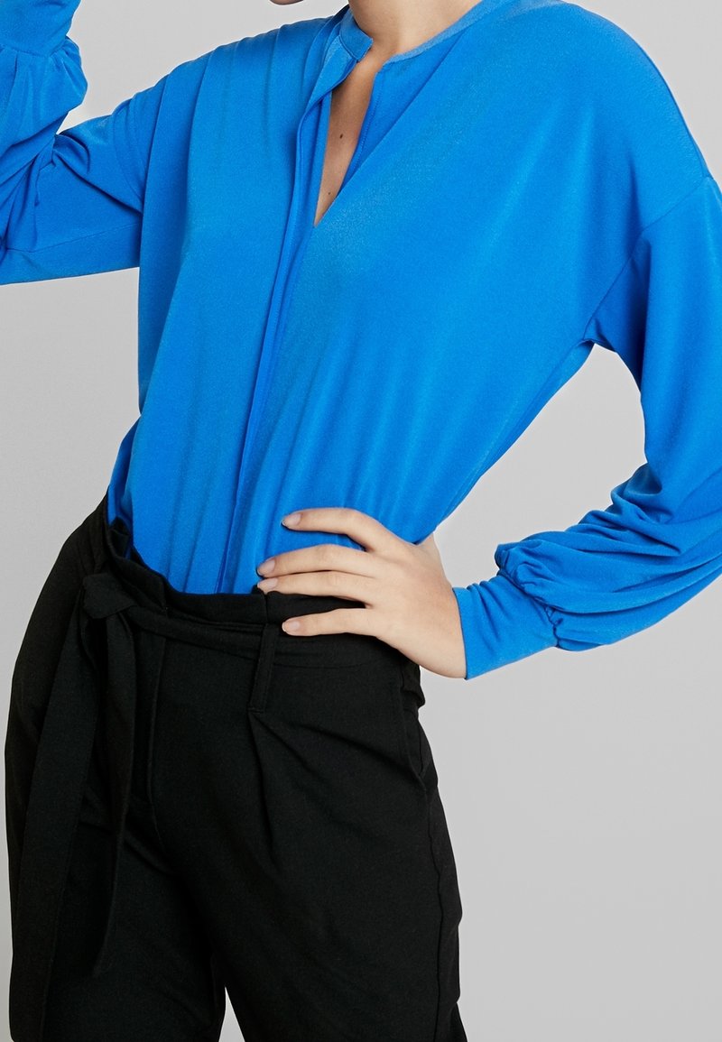 Långärmad blus i klarblått, med en delvis knäppning vid halsen. Tillsammans med svarta högmidjade byxor och ett knytbälte.