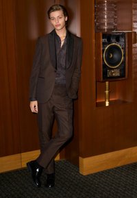 Braun gemusterter Anzug mit schwarzen Satin-Revers, kombiniert mit einem dunklen Paisley-Hemd. Glänzende schwarze Schuhe. Der Hintergrund zeigt Holzverkleidung und einen Lautsprecher.