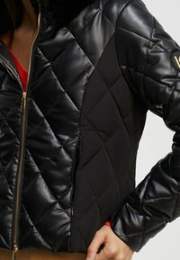 Svart vadderad bomberjacka med en glänsande yta, foder i päls vid kragen och dragkedja fram. Har sidofickor och en figursydd design.