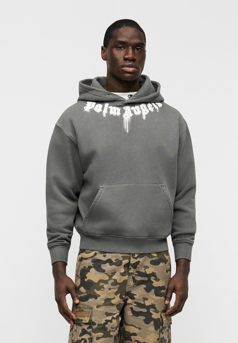 Γκρι φούτερ με κουκούλα με το λογότυπο "Palm Angels" σε λευκό χρώμα στο στήθος, τσέπη καγκουρό και ριμπ μανσέτες, σε συνδυασμό με καμουφλάζ παντελόνι cargo.