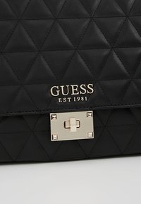 Sac à main en cuir matelassé noir avec logo GUESS doré et fermoir à verrou rectangulaire doré sur le rabat avant.