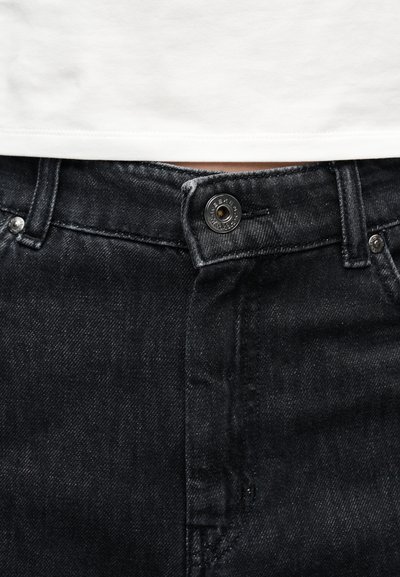 Κοντινή λήψη μαύρου τζιν παντελονιού με κουμπωμένο στη μέση, κάτω από λευκό πουκάμισο, που δείχνει θηλιές ζώνης και λεπτομέρειες ραφής.