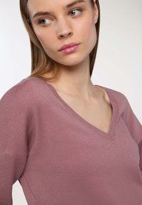 Maglione rosa chiaro a scollo a V con una texture morbida e bordi a coste. Presenta una vestibilità rilassata e maniche leggermente oversize.