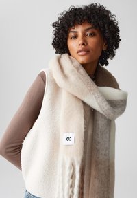 Scarf oversize beige e crema con una texture morbida e pelosa, drappeggiata attorno al collo, abbinata a un gilet bianco in pile con una patch del logo.