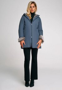 Cappotto grigio isolato con colletto e polsini in pelliccia sintetica, cappuccio, chiusura frontale con zip e due tasche laterali, abbinato a pantaloni neri aderenti.