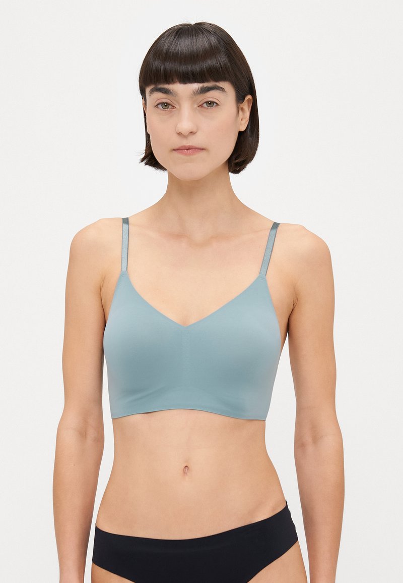 Helle blaue Bralette mit glatter Textur, V-Ausschnitt und verstellbaren Trägern. Getragen mit schwarzer Unterwäsche, vor einem weißen Hintergrund.