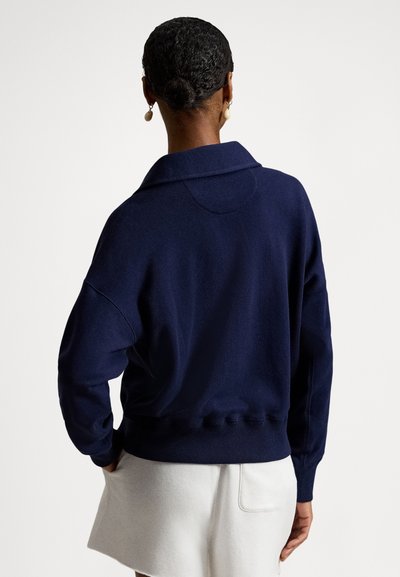 Γυναίκα με κοντά σγουρά μαλλιά σε χαμηλό σινιόν, φοράει χρυσά σκουλαρίκια κρεμαστά, μπλε πουλόβερ και λευκή φούστα, φαίνεται από πίσω μπροστά σε απλό φόντο.