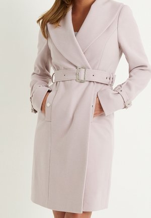 Kobieta nosząca jasnoróżowy płaszcz z paskiem, szerokim kołnierzem i zapinanymi na guziki paskami na mankietach i kieszeniach.