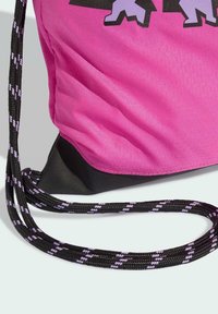 Pink drawstring bag with a textured surface, black bottom, and black-and-purple braided cord straps. Features graphic design.