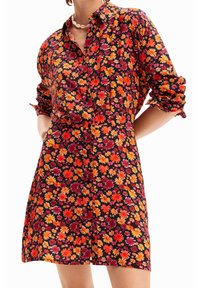 Sukienka koszulowa w kwiaty na ciemnym tle; zdobią ją pomarańczowe, czerwone i różowe kwiaty. Posiada zapięcie na guziki, podwinięte rękawy i luźny krój.