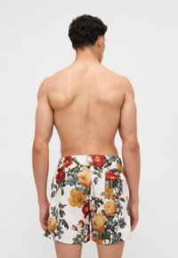 Costumi da bagno floreali con sfondo bianco, caratterizzati da grandi rose gialle e rosse, foglie verdi e due tasche posteriori.