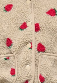 Krémovo sfarbená fleecová bunda s červenými vzormi jahôd, zapínaním na patentky vpredu a dvoma prednými vreckami.