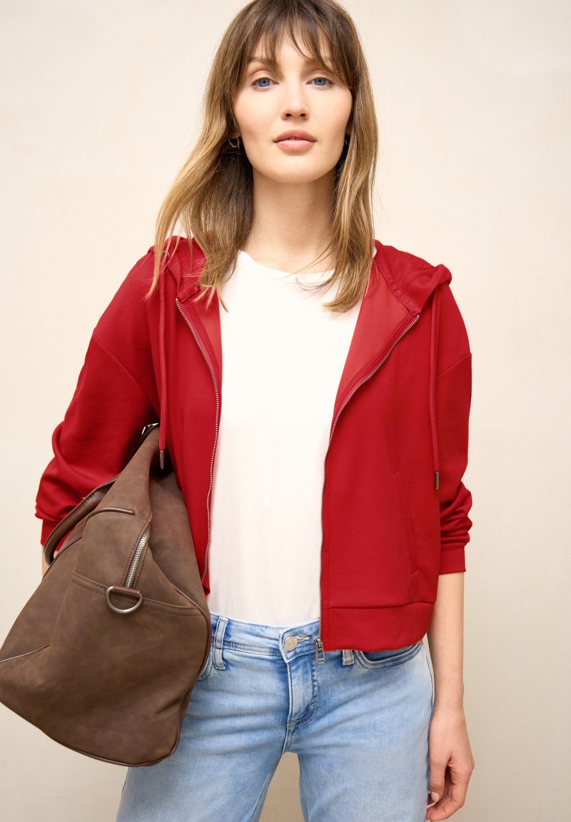 Jeune femme portant un sweat à capuche rouge zippé, un t-shirt blanc, un jean bleu clair, tenant un grand sac en daim marron sur l'épaule.