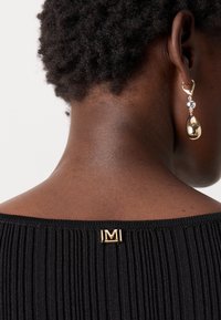 Top preto com textura, destacando um logo metálico "M" na gola, combinado com um brinco em forma de gota dourada com acentos prateados. Detalhe do tecido texturizado.