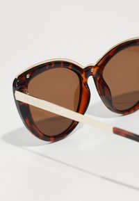 Óculos de sol estilo Gatinho em padrão tartaruga com lentes castanhas, detalhes dourados e uma haste de cor creme apresentando o texto "PRIVÉ REVAUX."