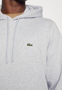 Grå bomullshuvtröja med en känguruficka, med en liten grön krokodil-logotyp på vänster bröst och snörningar i huva.