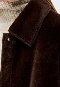 Manteau en fausse fourrure marron avec un grand col plat, doté d'une fermeture à bouton doré et d'une texture douce, porté par-dessus un pull en tricot de couleur claire.