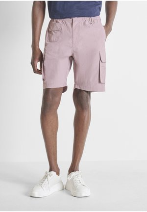 Modelo masculino con pantalones cortos cargo rosa claro con múltiples bolsillos y zapatillas blancas sobre un fondo liso.