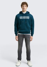 Teal hoodie met een logo op de voorkant, een buidelzak en een koord. Gecombineerd met grijze jeans en witte sneakers, in een casual uitstraling.