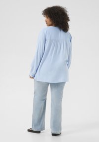 Lichtblauwe blouse met lange mouwen en een geplooide achterkant, gecombineerd met lichtblauwe wijde spijkerbroek en zwarte platte schoenen. Glad stoftextuur.