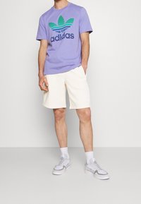 Lila T-shirt med grön och blå randig logotyp, i kombination med krämfärgade shorts. Inkluderar vita sneakers och vita strumpor. Enkel design och passform.