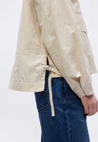 Μπεζ μακρυμάνικο πουκάμισο με πλαϊνή σχισμή και δεμένη ταινία, φορεμένο με μπλε τζιν παντελόνι στο κάτω μέρος του κορμού και το χέρι ενός ατόμου.