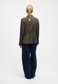 Blazer marrone su misura con silhouette strutturata e due bottoni posteriori, abbinato a pantaloni blu navy dalla vestibilità ampia. Tessuto liscio e strutturato.