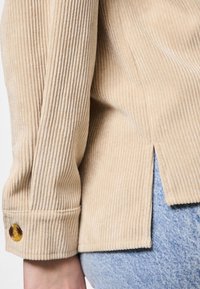 Beige väriyksityiskohtainen puuvillainen corduroy-paita, jossa pystysuora ribbineulos, sivuhalkiot ja ruskea nappi hihansuussa. Päällä vaaleansiniset farkut.