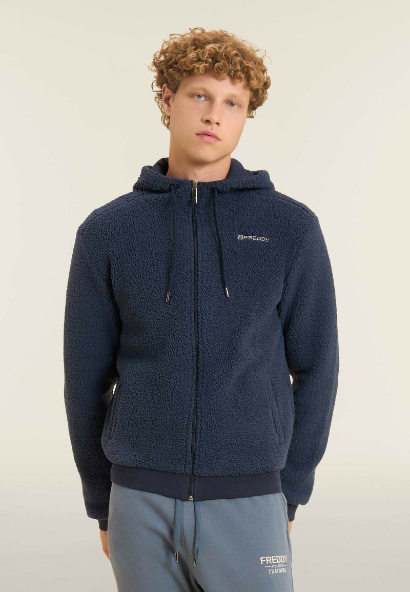 Chaqueta de forro polar azul marino con capucha, cierre de cremallera y dos bolsillos laterales. Textura suave con una cinturilla y puños elásticos. Logo de Freddy en el pecho.