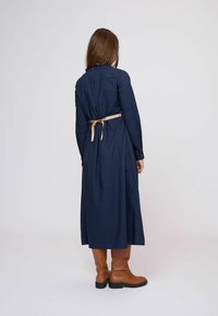 Tengerészkék hosszú ruha hosszú ujjakkal, derékrészen világos bézs szalaggal megkötve, sima anyagból készült. Modell barna térdig érő csizmát visel.