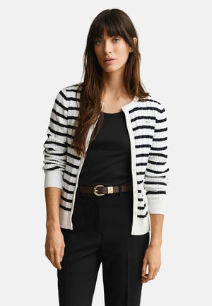 Vrouw met lang bruin haar draagt een zwart topje, een wit-zwarte gestreepte cardigan, zwarte broek en een bruine riem, staand tegen een effen achtergrond.