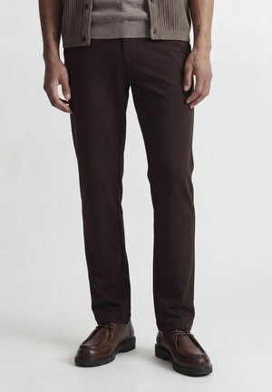 Mand iført mørkebrune bukser, brune læder snørestøvler og en beige knappet cardigan mod en ensfarvet baggrund.