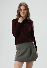 Bordeaux gebreide polotrui met een ruitenpatroon, voorzien van een knopenlijst. Gecombineerd met een grijze, op maat gemaakte korte rok met zijaccenten.
