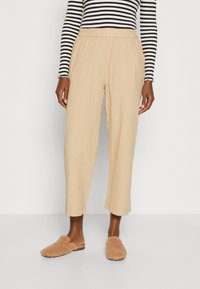 Pantalons beiges, légers, à taille élastique avec une coupe décontractée et des jambes fuselées. Associés à des chaussures à enfiler en peluche de couleur tan. Un haut à rayures à manches longues est visible.